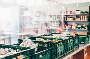 Lebensmittelkisten stehen hinter einer Trennwand, eine Tafel-Mitarbeiterin sortiert Lebensmittel