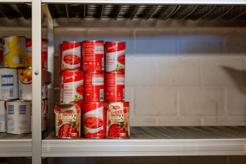 Blick in ein Edelstahlregal, das nur zu einem Drittel mit Tomaten-Konversendosen gefüllt ist.