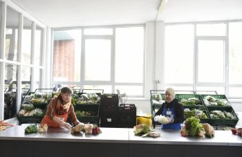 Lebensmittelkisten stehen hinter einer Trennwand, eine Tafel-Mitarbeiterin sortiert Lebensmittel