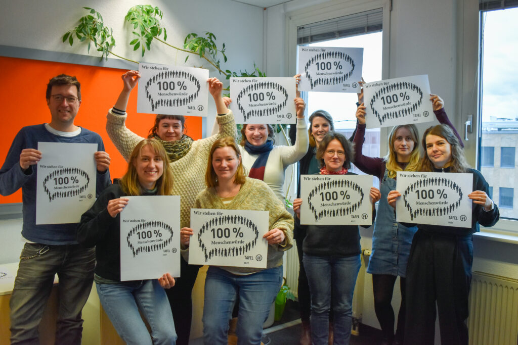 Tafel-Aktive lächeln in die Kamera. In den Händen halten sie Schilder mit der Aufschrift "Wir stehen für 100 % Menschenwürde"