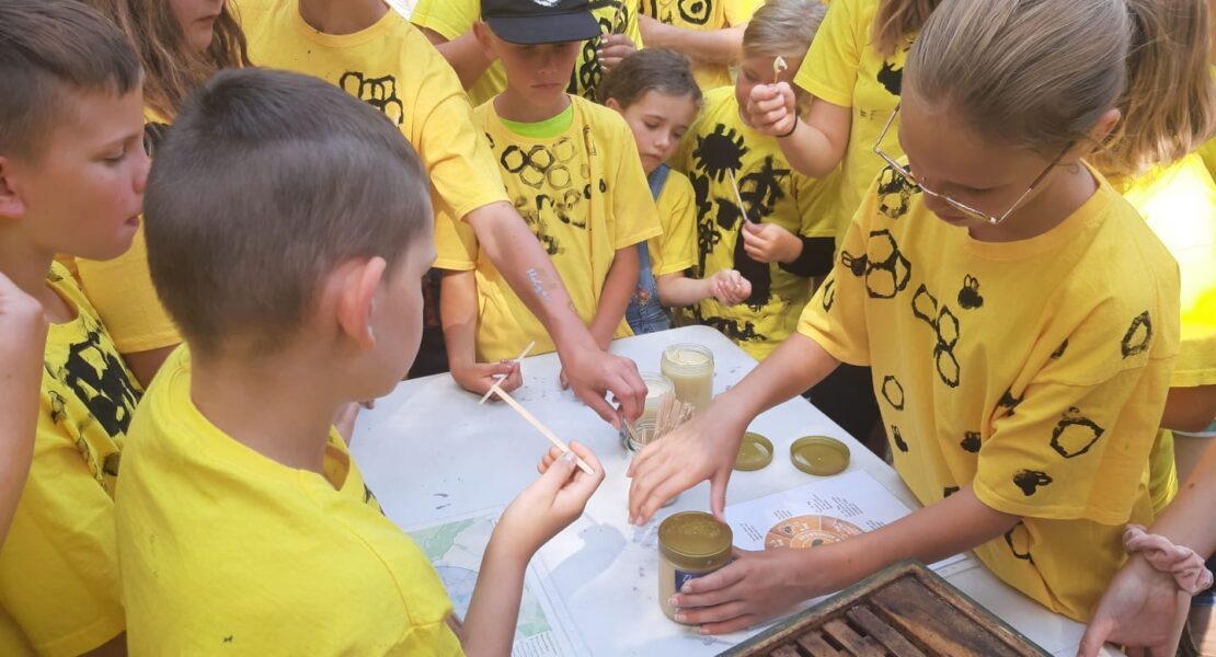 Kinder in mit Bienen bemalten gelben T-Shirts stehen gemeinsam um einen Tisch mit Honiggläsern