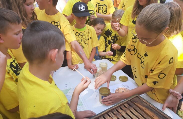 Kinder in mit Bienen bemalten gelben T-Shirts stehen gemeinsam um einen Tisch mit Honiggläsern