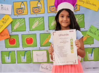 Junges Mädchen mit Kochmütze auf dem Kopf und Urkunde in der Hand vor einer Ernährungspyramide