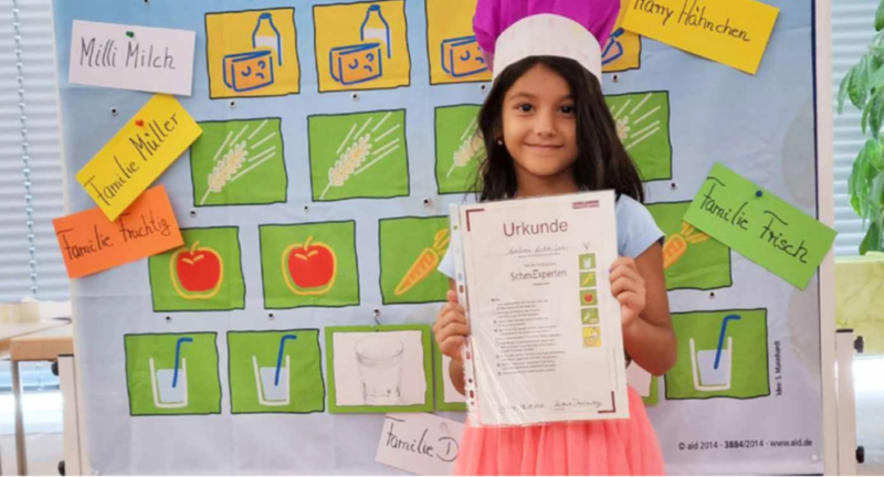 Junges Mädchen mit Kochmütze auf dem Kopf und Urkunde in der Hand vor einer Ernährungspyramide