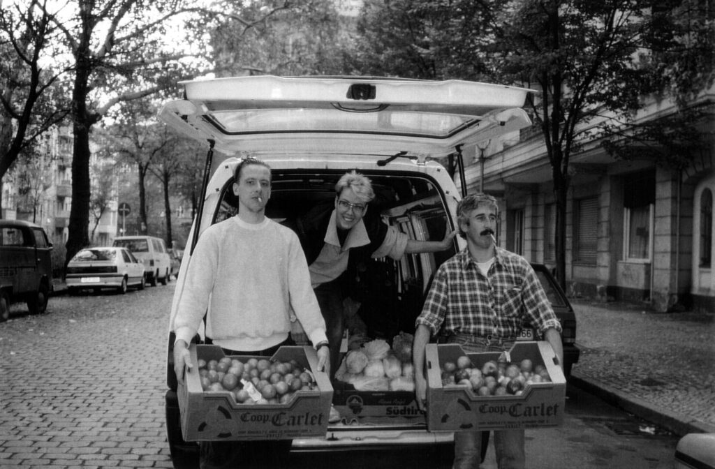 Schwarz-Weiß-Foto von Sabine Werth, die im Laderaum eines Autos hockt. Davor stehen zwei Tafel-Helfer, die jeweils eine Kiste mit Obst tragen.