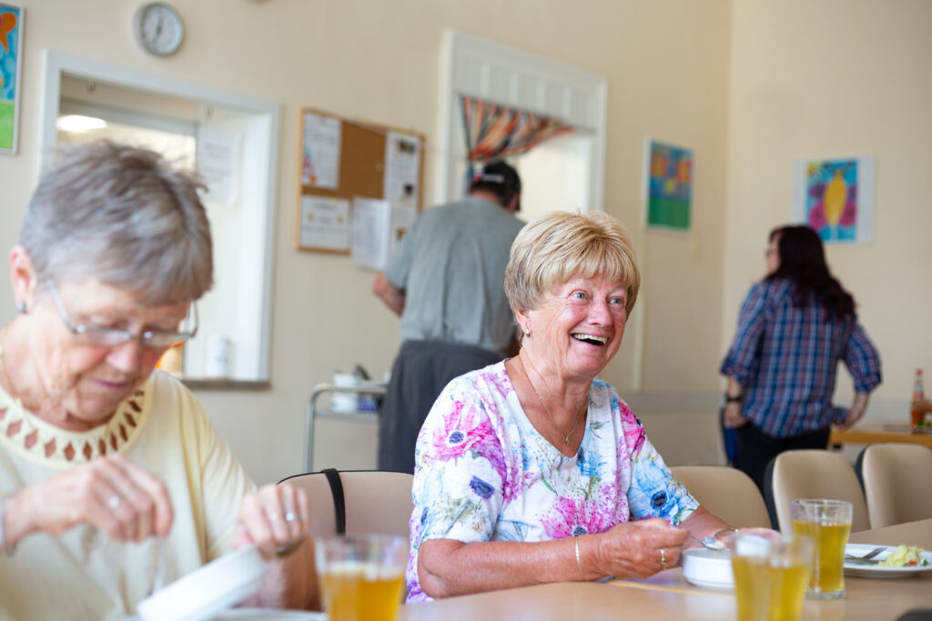 Zwei ältere Frauen sitzen lachend am Tisch und essen gemeinsam