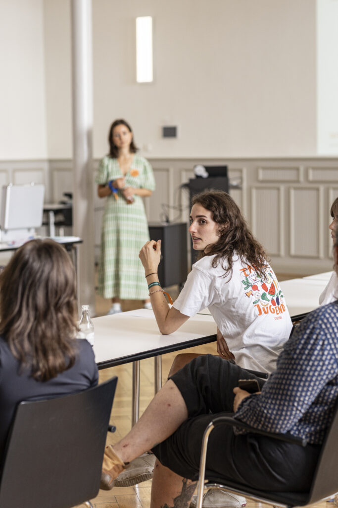 2 junge Frauen sitzen an einem Tisch und sprechen miteinander. Im Hintergrund steht die Workshop-Leiterin und hört ihnen zu.