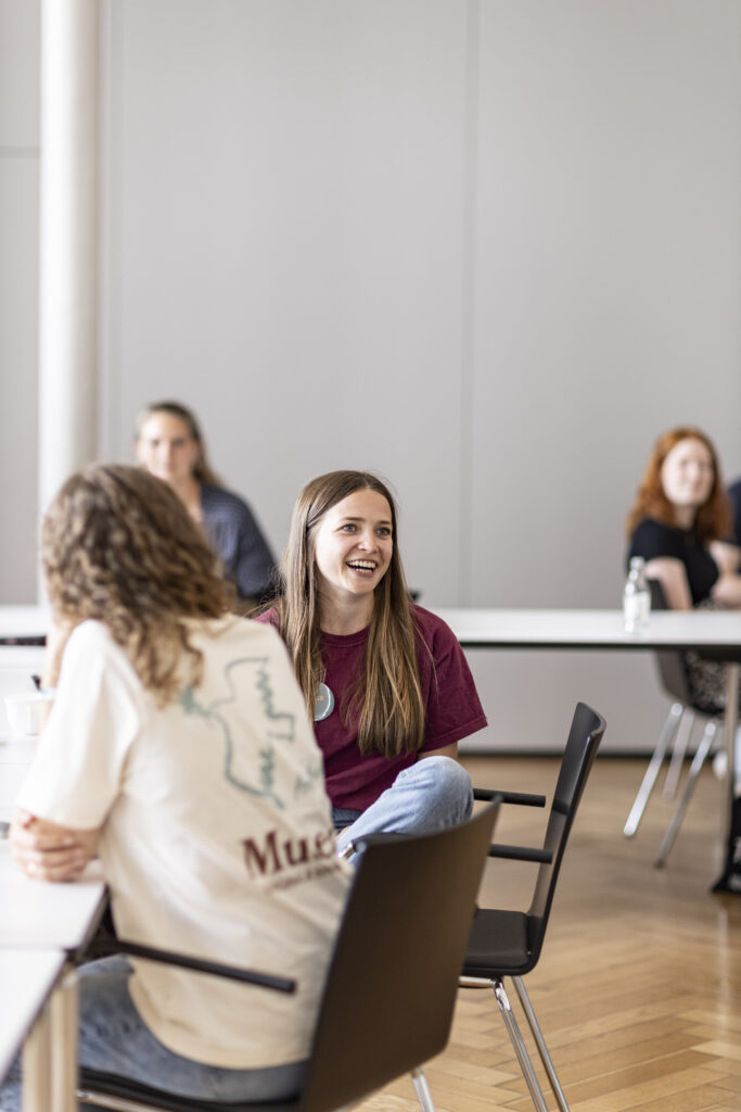 Eine junge Ehrenamtliche sitzt an einem Tisch in einem Workshop und spricht.