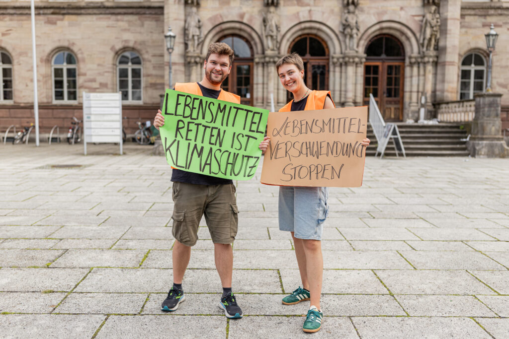 2 Tafel-Helfer:innen posieren in Göttingen mit Plakaten gegen Lebensmittelverschwendung