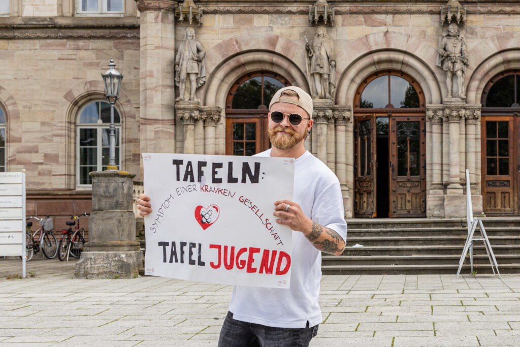 Ein Tafel-Aktiver posiert in Göttingen mit einem Plakat, auf dem steht "Tafeln: Symptom einer kranken Gesellschaft"