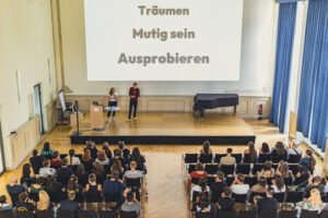 Auf der Bühne beim Tafel Jugend-Kongress 2024 stehen 2 Personen, auf die wir schräg von oben blicken. Vor ihnen sitzt das Publikum und auf die Wand sind die Wörter "Träumen, Mutig sein, Ausprobieren" projiziert.