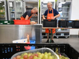 Zwei Tafel-Helfer sortieren Kühlware in Kühlschränke ein. Im Vordergrund sind Weintrauben zu sehen.