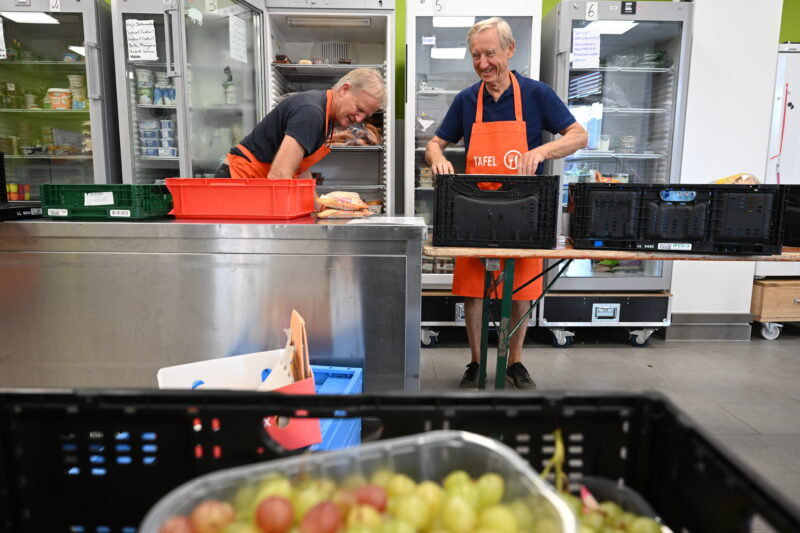 Zwei Tafel-Helfer sortieren Kühlware in Kühlschränke ein. Im Vordergrund sind Weintrauben zu sehen.