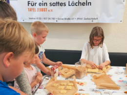 Kinder dekorieren konzentriert Plätzchen. Im Hintergrund ist zu lesen: "Für ein sattes Lächeln - Tafel Zerbst"