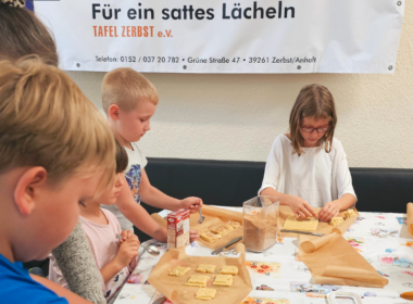 Kinder dekorieren konzentriert Plätzchen. Im Hintergrund ist zu lesen: "Für ein sattes Lächeln - Tafel Zerbst"