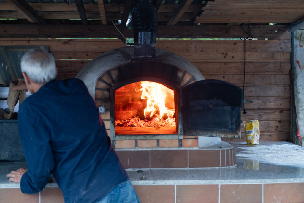 Das Feuer im Holzbackofen brennt, ein Mann steht davor. 
