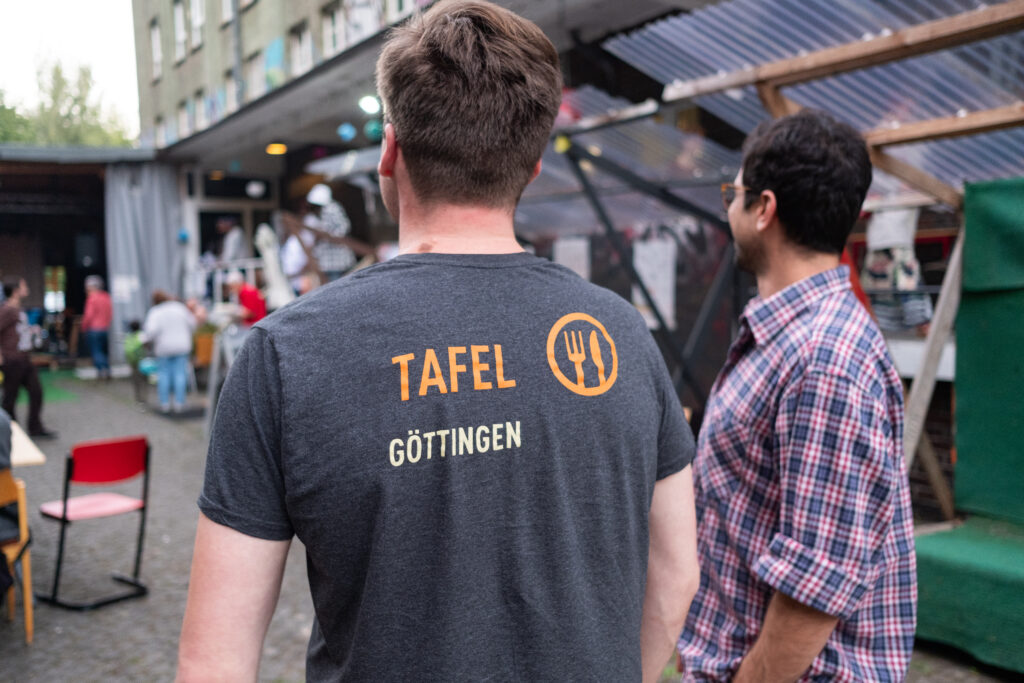 Zwei Männer schauen auf die Bühne und das Gelände. Einer trägt ein T-Shirt mit der Aufschrift Tafel Göttingen. 