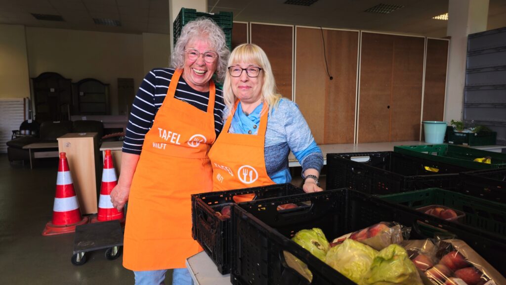 Zwei Tafel-Helferinnen, die orangefarbene "Tafel hilft"-Schürzen tragen,lächeln in die Kamera