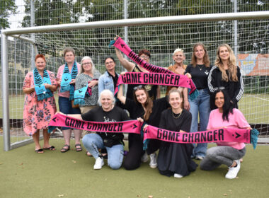 12 Frauen stehen in einem Tor und lächeln in die Kamera. Sie halten Schals mit der Aufschrift "Game Changer".