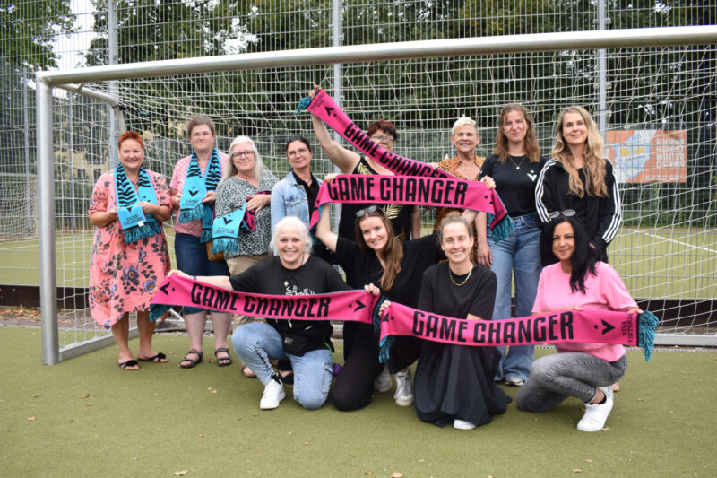 12 Frauen stehen in einem Tor und lächeln in die Kamera. Sie halten Schals mit der Aufschrift "Game Changer".