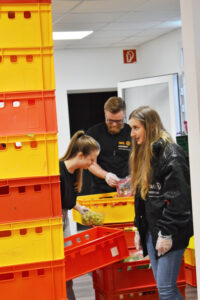 3 Tafel-Helfer:innen sortieren Lebensmittel neben einem Stapel leerer Kisten.