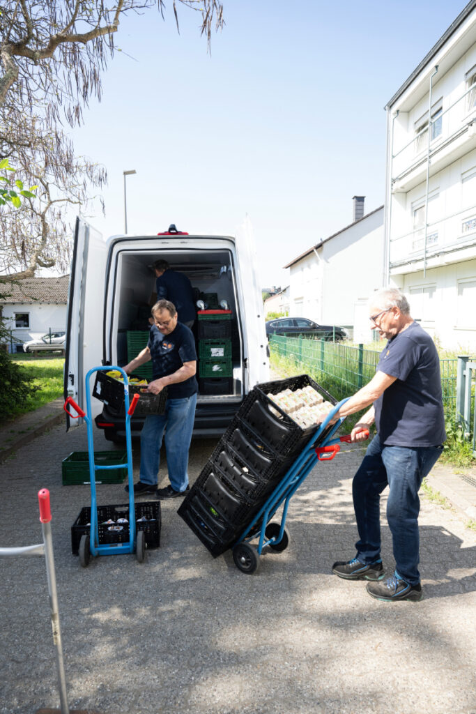 2 Tafel-Helfer laden Kisten voller Lebensmittel aus einem Fahrzeug aus.
