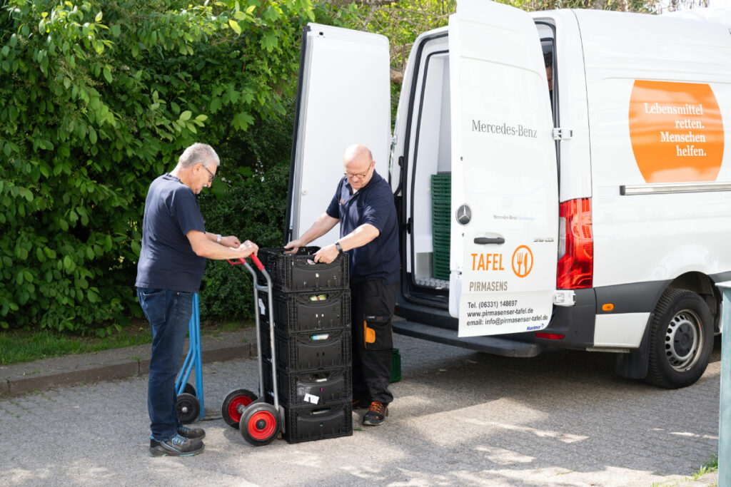 2 Tafel-Helfer beladen eine Sackkarre mit vollen Lebensmittelkisten.