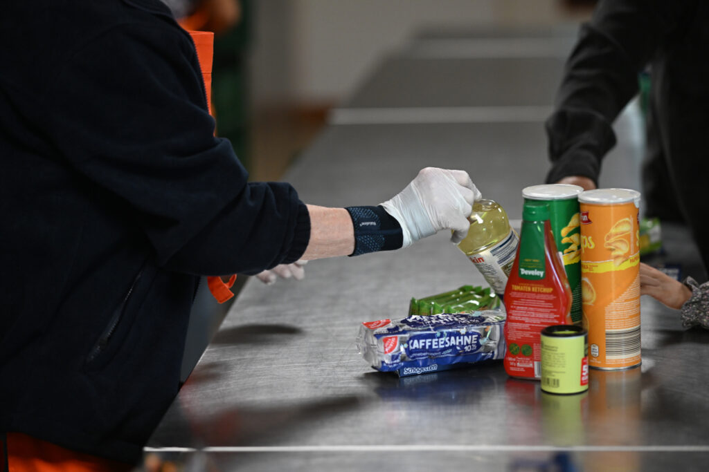 Eine Tafel-Helferin stellt einem Kunden verschiedene haltbare Lebensmittel auf die Theke, darunter Öl, Chips und Ketchup.