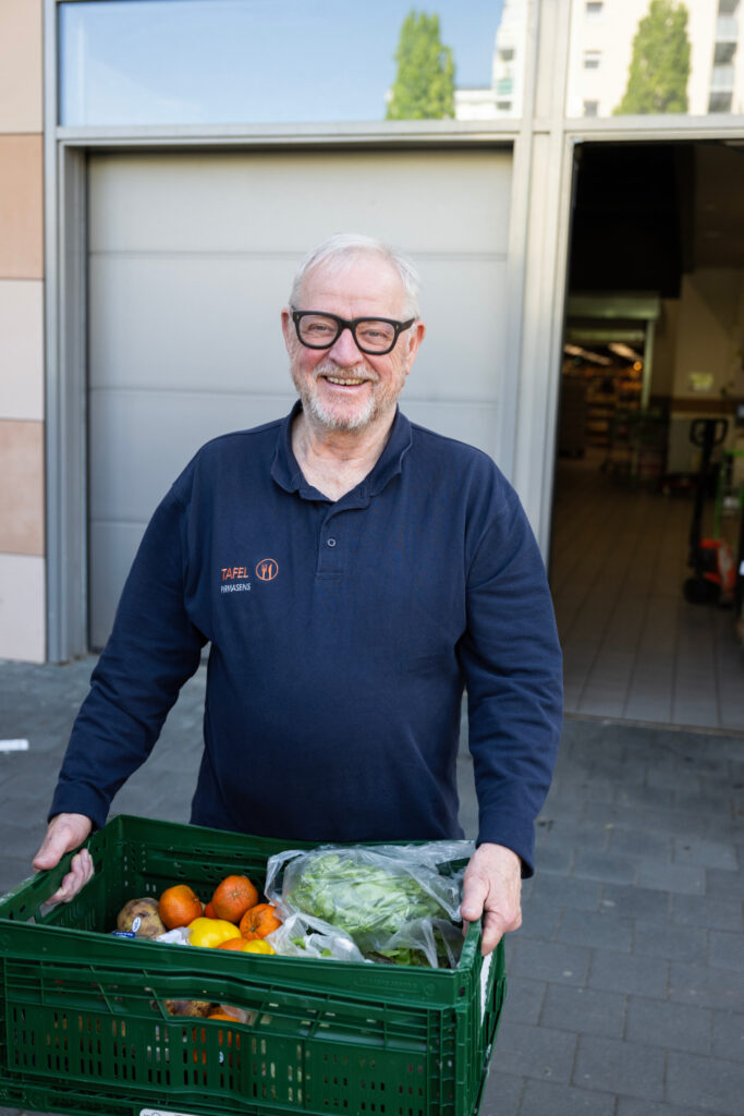 Wolfgang Nikolaus, Vorsitzender Pirmasenser Tafel, trägt lächelnd eine Kiste mit Obst und Gemüse